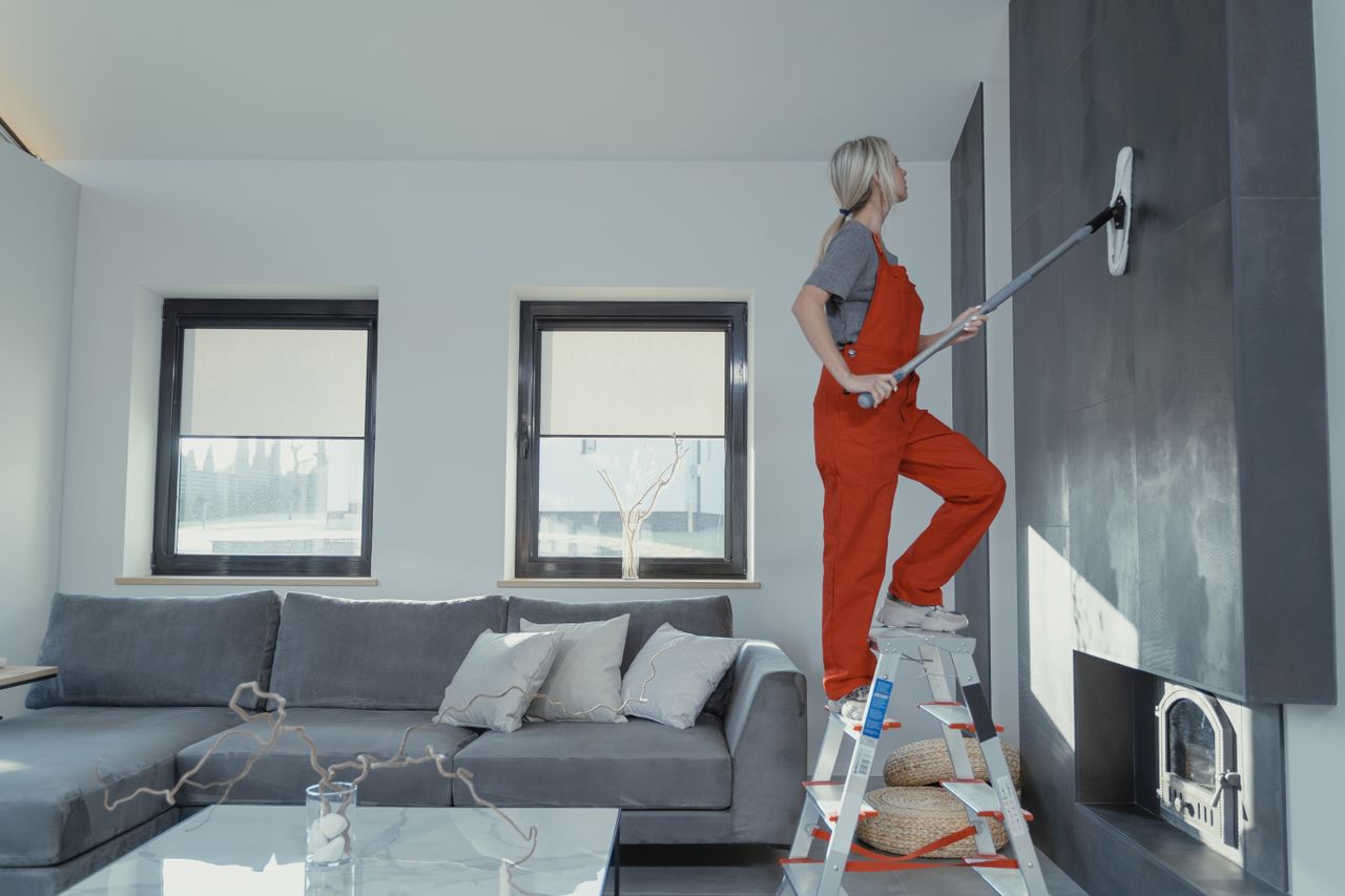 team-02 A woman in overalls cleans a wall in a stylish living room, enhancing home hygiene.